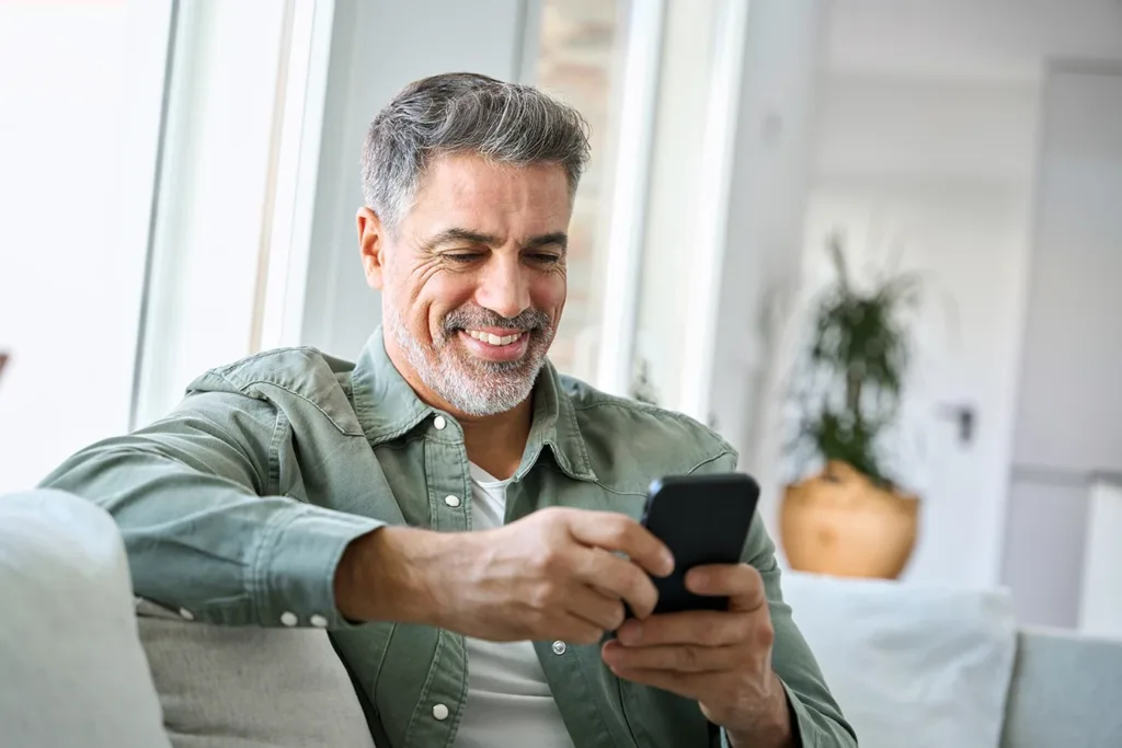 A mature man, with gray hair and a salt and pepper beard, sitting on his couch, looking down at his smartphone and smiling, symbolizing New Hearing Aid Connectivity Technology in 2026
